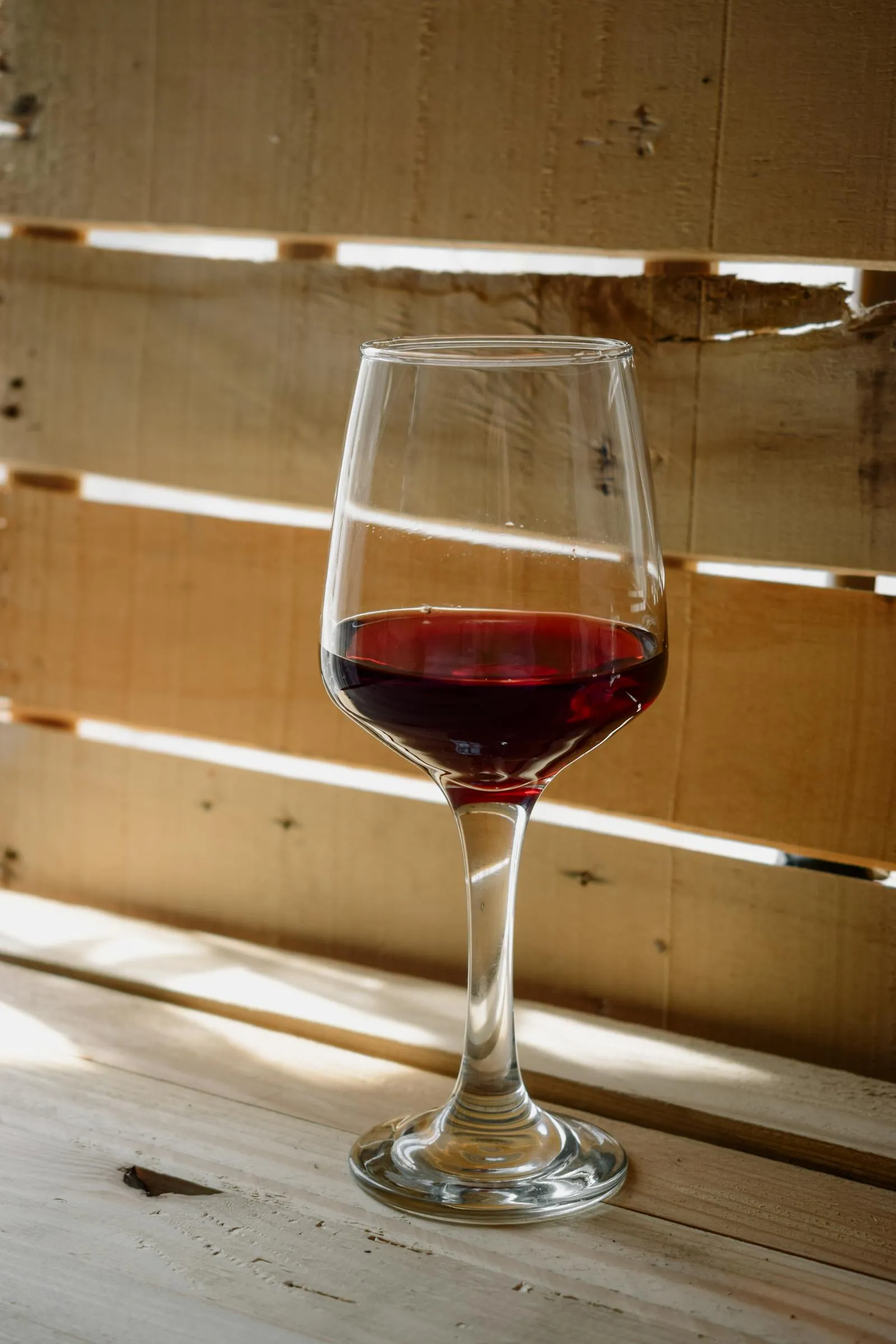 A single glass of red wine stands on a rustic wooden surface in natural light